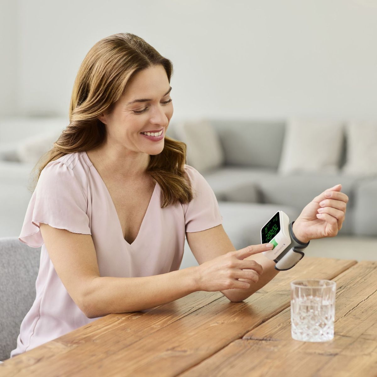 Eine lächelnde Frau sitzt an einem Holztisch und misst ihren Blutdruck mit einem digitalen Handgelenk-Blutdruckmessgerät, während ein Glas Wasser vor ihr steht.